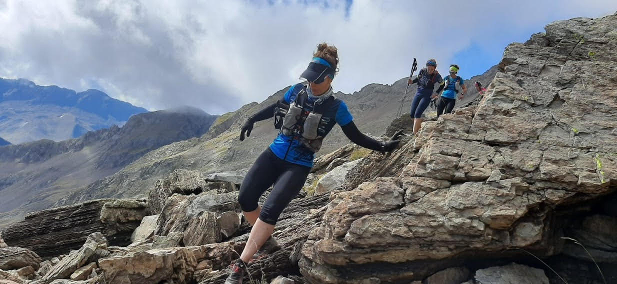 Descripción de la foto de portada: En primer plano se ve mujer realizando una bajada corriendo por zona pedregosa. Tras ella, algo más alejadas se ven otras tres mujeres. De fondo todo un paisaje de montañas con cielo cubierto de nubes