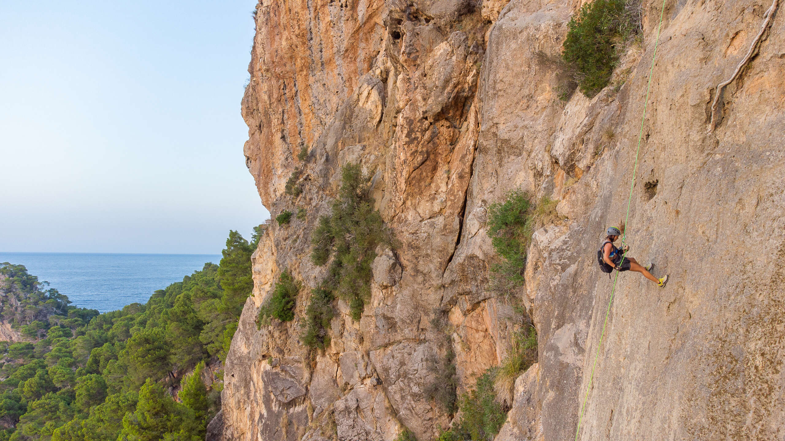 Descripción de la foto de portada: En primer plano aparece una mujer rapelando la pared de una montaña con cuerda verde, casco y mochila. Al fondo se ve un mar azul y entre la roca y el mar un manto verde de árboles.