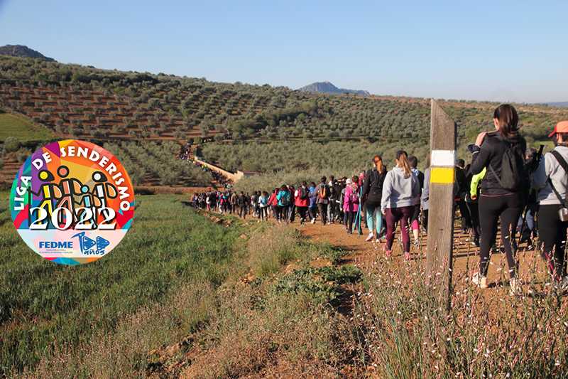 Foto de portada Fotografía en color de un grupo de personas caminando en un paisaje dominado por los olivos; en primer término, aparece una estaca con una señal de PR®. Sobre la misma aparece el logo de la Liga de Senderismo de la FEDME.