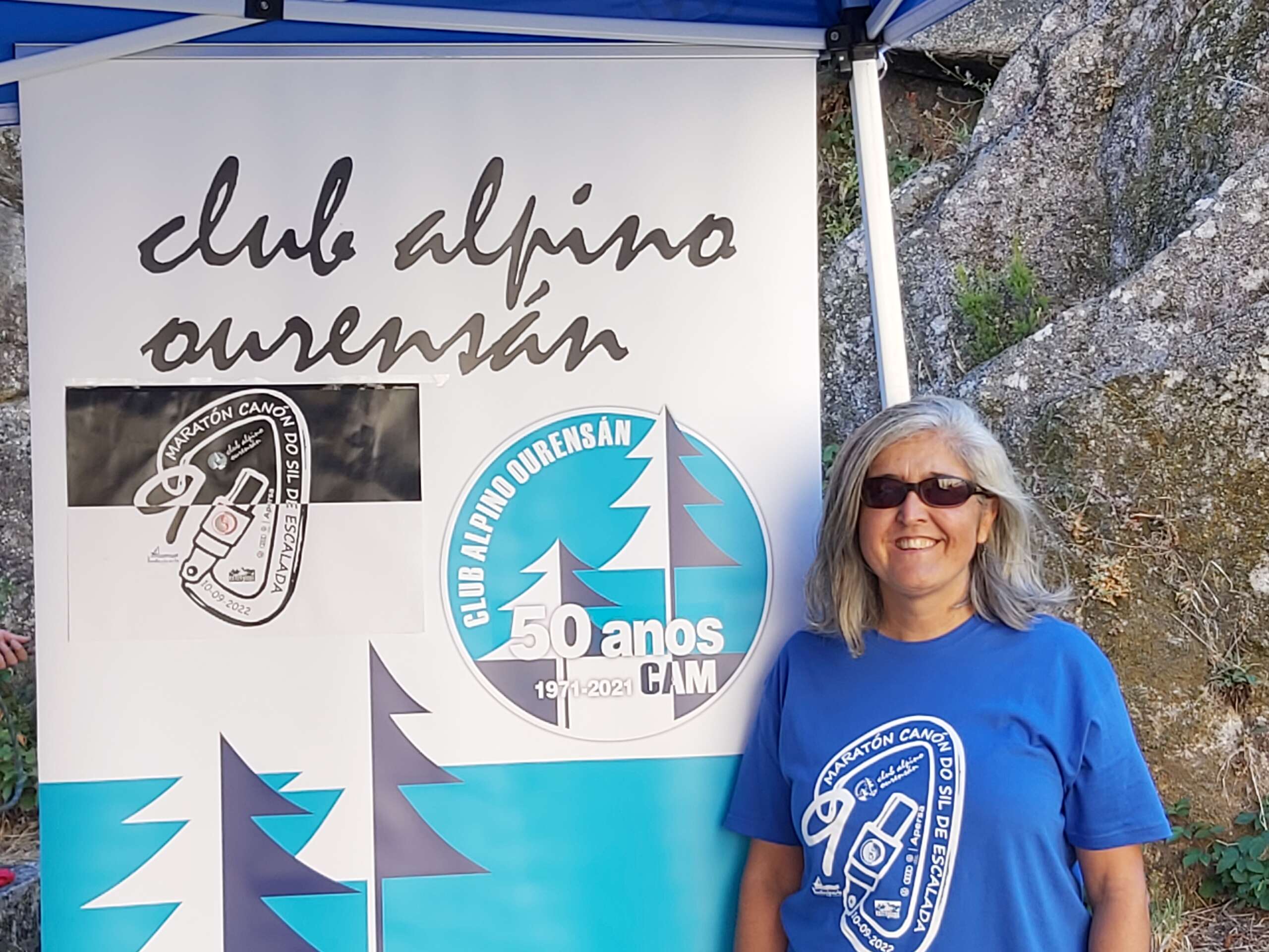 Descripción foto de portada: Mar Pallás Valleros aparece sonriente en lado derecho de la foto con camiseta azul con un estampado del logo de la Maratón Cañón Do Sil de Escalada, es un mosquetón abierto. A su lado ocupando gran parte de la foto el cartel anunciador de dicho evento con el nombre de su Club Alpino Ourensán arriba y debajo los logos del maratón ya descrito y el de dicho club que es redondo, con dos pinos uno más pequeño que el otro en blanco y gris y debajo pone: 50 años (1971-2021)