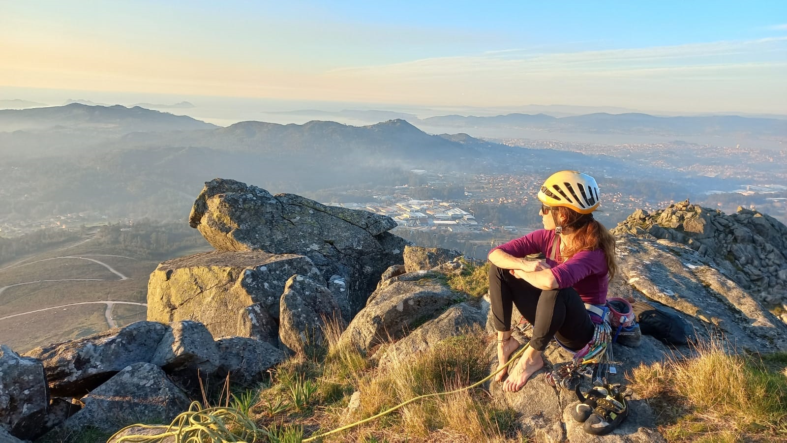 Foto de portada: Katya Sanz Ochoa aparece en la cumbre del Monte Galiñeiro, sentada en una roca con las piernas recogidas y sujetas por sus brazos. Mirada hacia un lado donde se ve de fondo unos valles, pueblos y montañas. Ella lleva arnés, material de escalada, cuerda y casco.