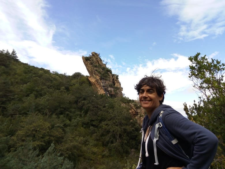 Inés Bállega aparece en primer plano de cintura para arriba, con chaqueta y mochila azul a la espalda y muy sonriente. Tras ella un bosque de árboles y una gran roca emergiendo entre ellos