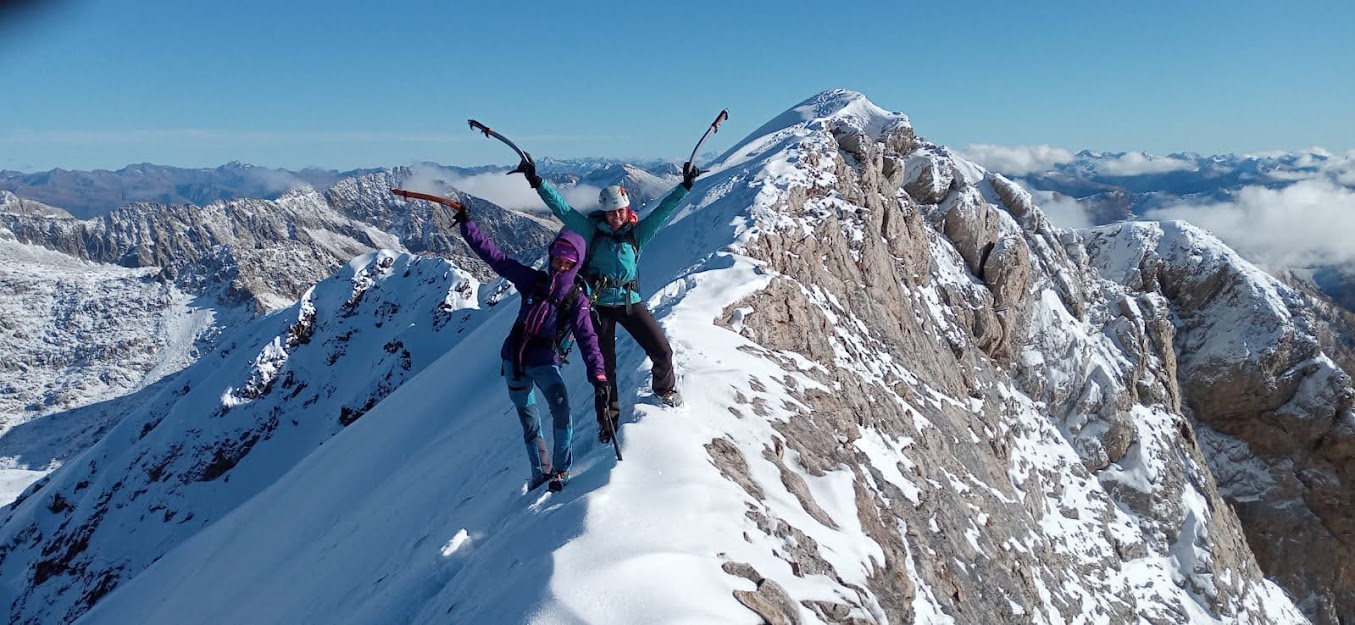 Dos mujeres alpinistas aparecen en la cresta de la cumbre de una montaña con nieve y roca. Ellas con equipamiento de alta