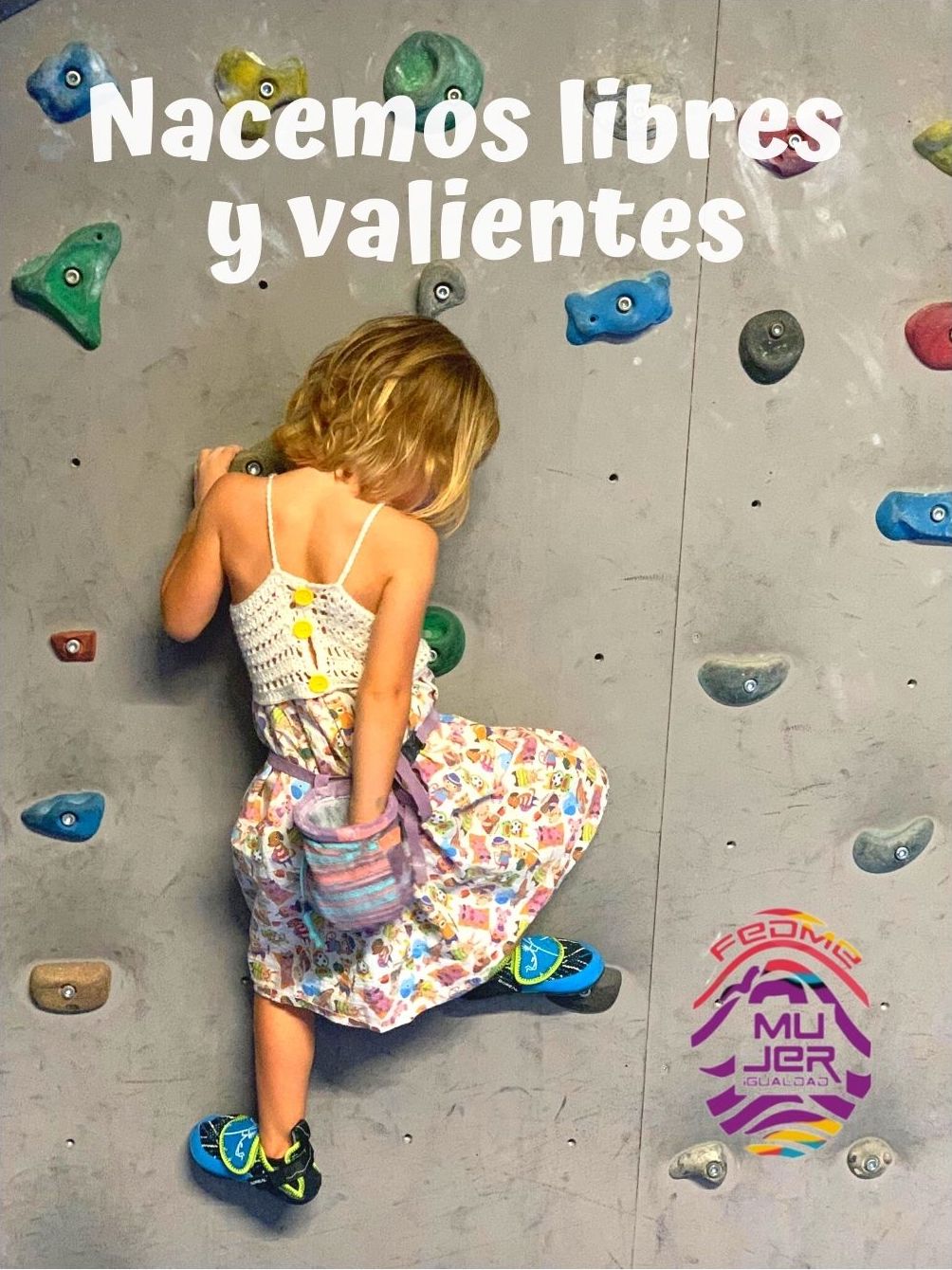 En una pared de rocódromo una niña pequeña, de unos 4 años, escala con gran estilo y tranquilidad. Se la ve con la mano izquierda agarrada a una presa y con la derecha impregnando su mano de polvo de magnesio en la bolsa que le cuelga por detrás. Luce un vestido de niña, con la parte superior tejida de ganchillo y la falda de dibujos. En los pies lleva sus correspondientes gatos y se apoya con ellos en dos presas. Todo bajo el lema “NACEMOS LIBRES Y VALIENTES”