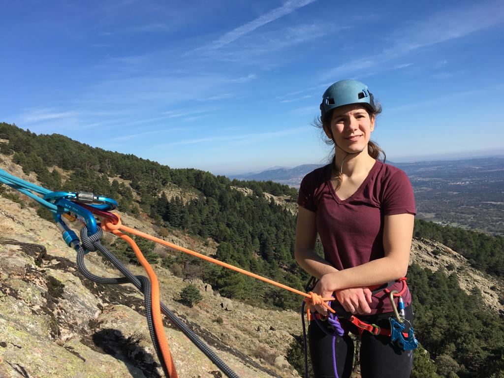 Andrea sale a la derecha de la foto en una reunión con arnés y casco de escalada, muy sonriente. Está en una pared de piedra en El Escorial y detrás se ve la ladera de la montaña.