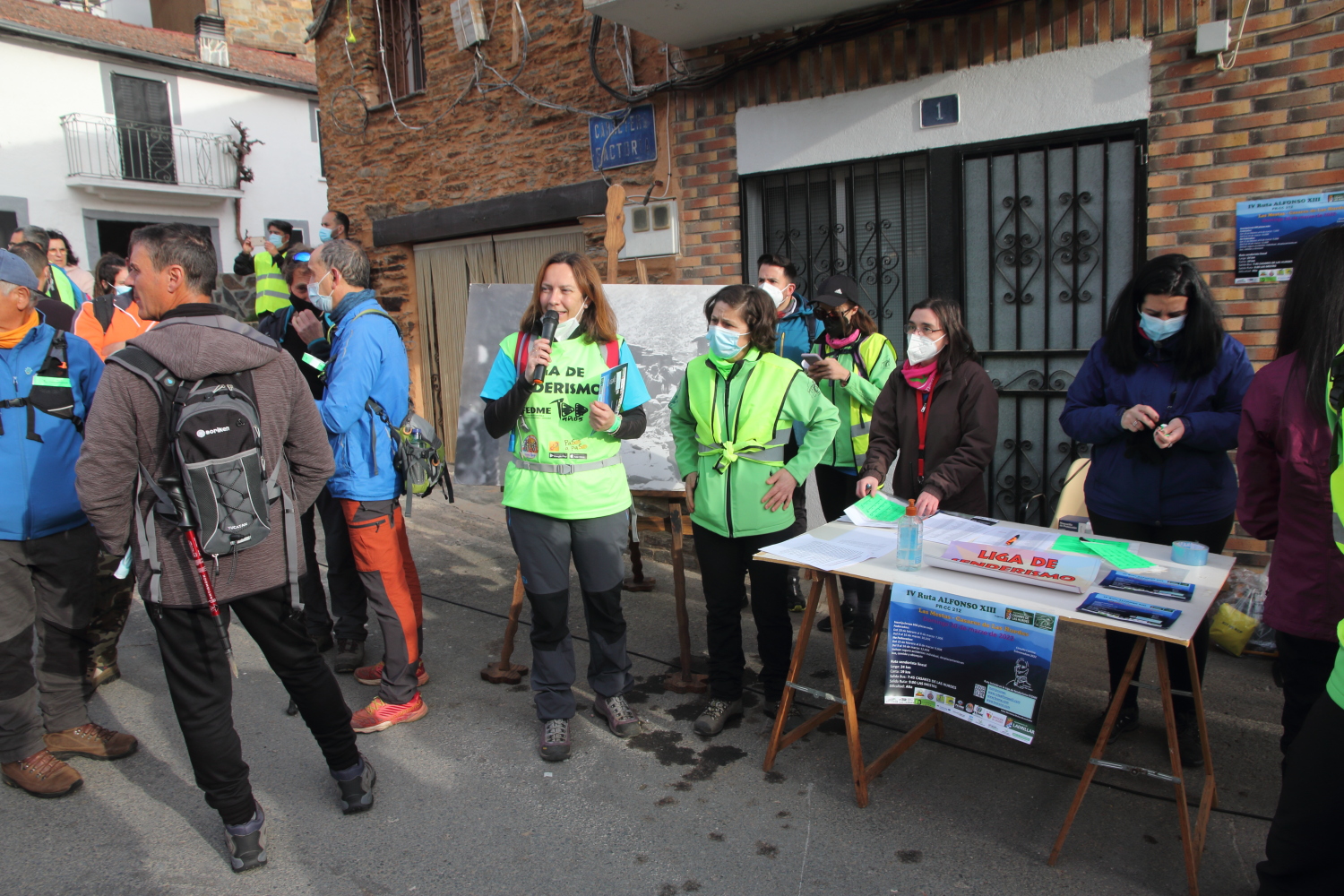 Fotografía que ilustra el momento del registro y explicaciones de la IV ruta de Alfonso XIII. Pequeño grupo de personas senderistas en torno a la mesa de inscripciones, al inicio de la ruta. Aparece en el centro, de pie, la persona organizadora, con el peto de Liga de Senderismo, explicando los pormenores de la actividad con el uso de un micrófono