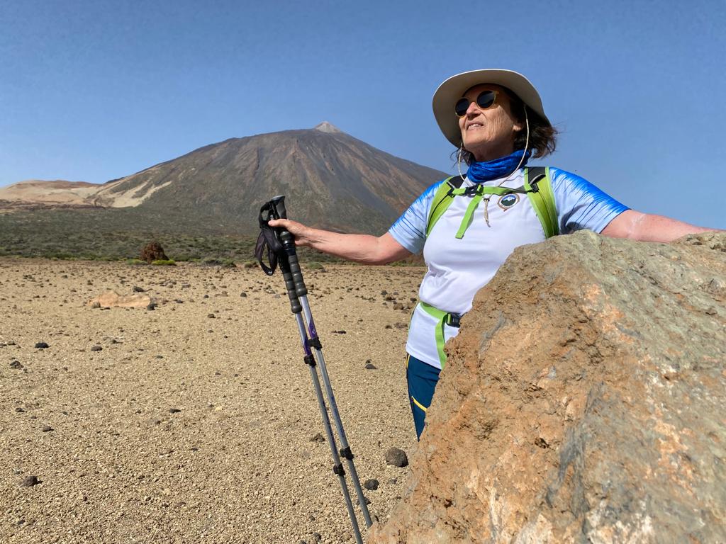 Josefina Suárez aparece en primer plano tras una roca en Parque Nacional del Teide. Su brazo izquierdo apoyado sobre la roca y el derecho con dos bastones hacia el suelo. Ella equipada con ropa de montaña, sombrero y gafas y mirando sonriente hacia arriba y al frente. Tras ella un gran llano de arena blanca y en el fondo el imponente Teide
