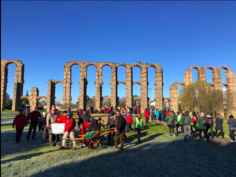 Fotografía en color que presenta el momento en el que los participantes en la XIV Ruta Senderista Monumental Octavio Augusto, integrada en la Liga Ibérica de Senderismo, andan junto al acueducto romano de Los Milagros. En primer plano aparece un grupo llevando a una persona con discapacidad en silla joelette, mientras que el resto del senderistas se dirige para pasar entre los arcos del acueducto.