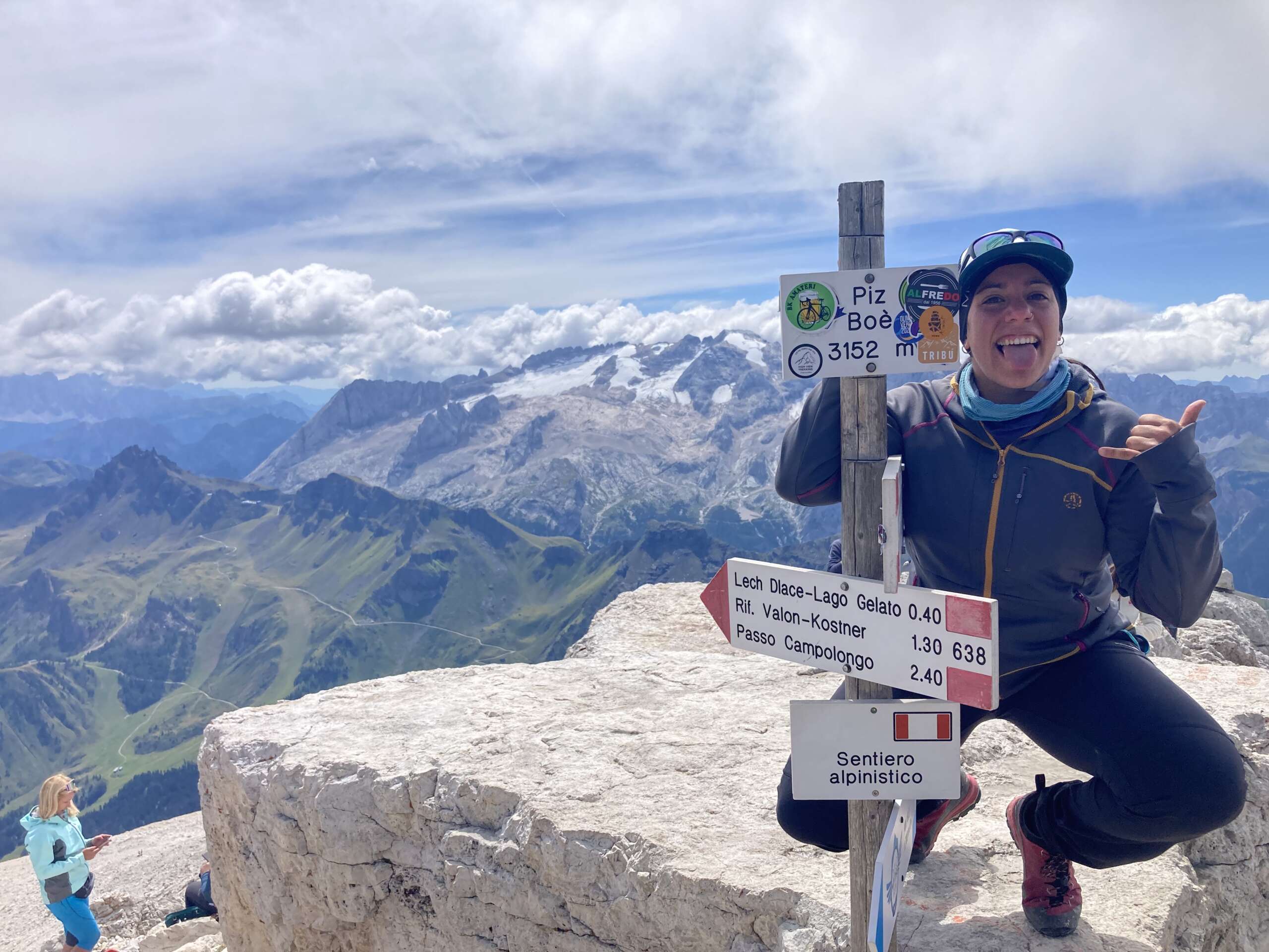Laura Martín aparece en primer plano en cuclillas, encima de una gran roca de la cumbre del Piz Boè de 3152 metros, apoyada con señal delante de ella donde se marca dicho pico. Se la ve feliz y sacando la lengua. Al fondo un bonito paisaje de montañas, algunas con nieve, nubes y cielo azul