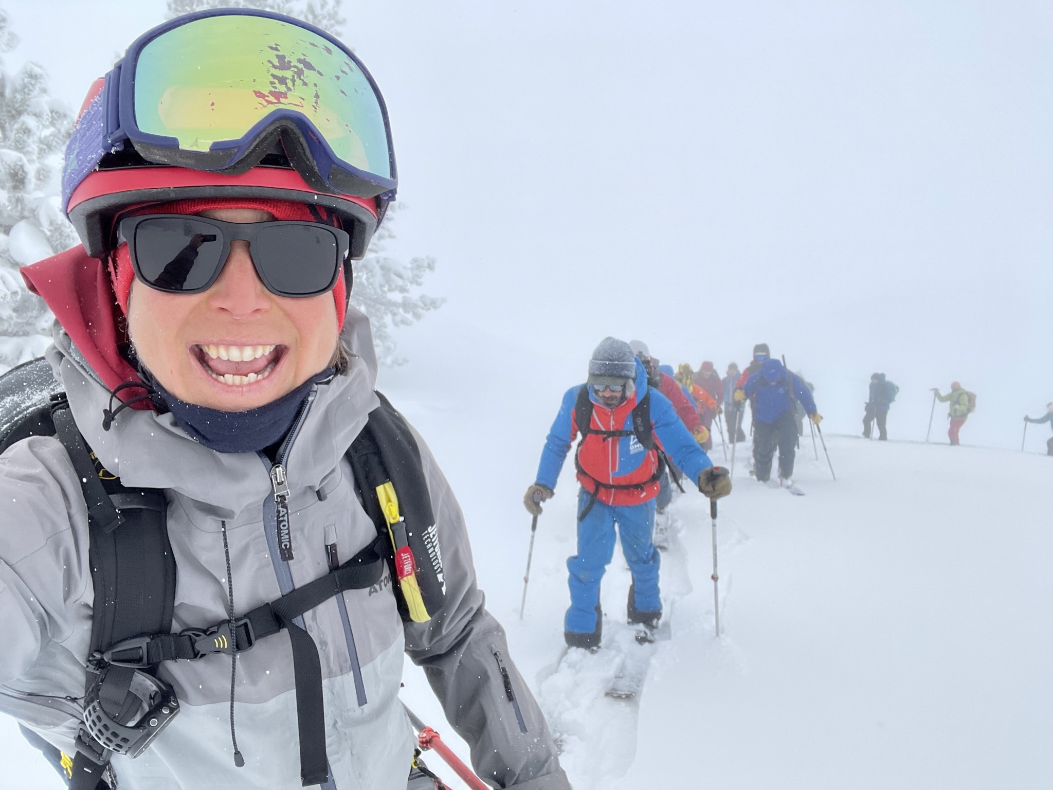 Montse aparece a la izquierda de la foto con indumentaria invernal en un entorno totalmente nevado. Está realizando un selfie por lo que se la ve en primer plano. Detrás y progresando en condiciones rigurosas y preciosas de pleno invierno hay un grupo de instructores ACNA para realizar una práctica de rescate en avalancha. La foto fue tomada en Orri, Val d'Aran en enero de 2023.