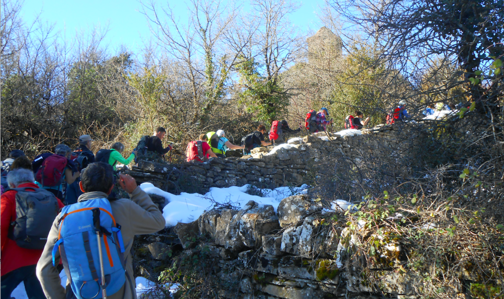 Fotografía en color que presenta a un grupo de senderistas avanzando por un camino tradicional; al fondo aparece la silueta de la torre de una iglesia