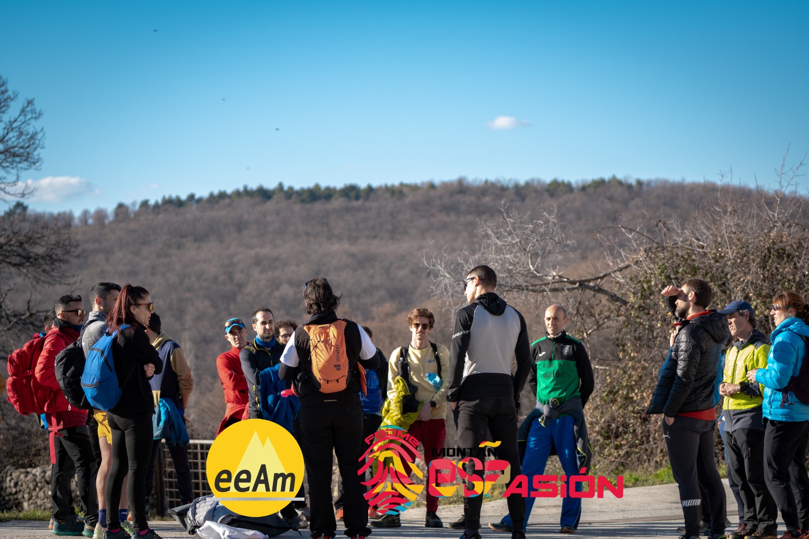 en la foto aparece un grupo de aproximadamente 20 personas, todas ellas vestidas con equipación deportiva, colocados en círculo y atendiendo las explicaciones de una persona del grupo. El grupo está situado en un entorno natural, ya que al fondo se puede observar una montaña de poca altitud.