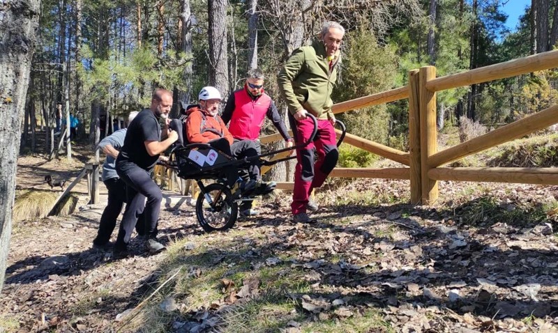 Grupo de alumnos practicando con una silla Joëlette suben una pendiente ligera por terreno irregular.