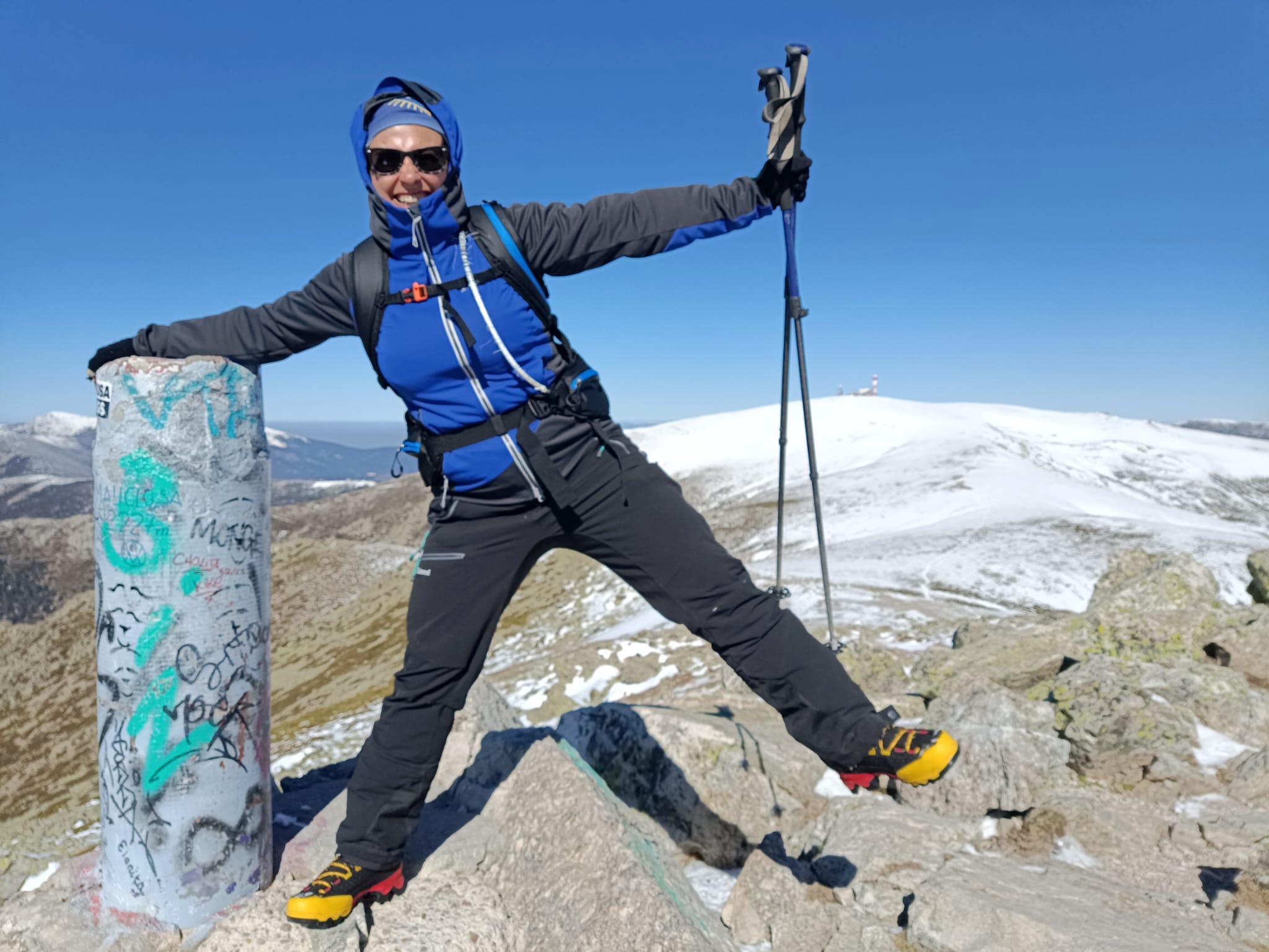 Henar está en el Pico de la Maliciosa en Madrid posando apoyada en el vértice geodésico con una mano y abriendo la pierna contraria en el aire. Viste ropa deportiva de invierno y porta mochila y unos bastones. Al fondo una loma nevada.