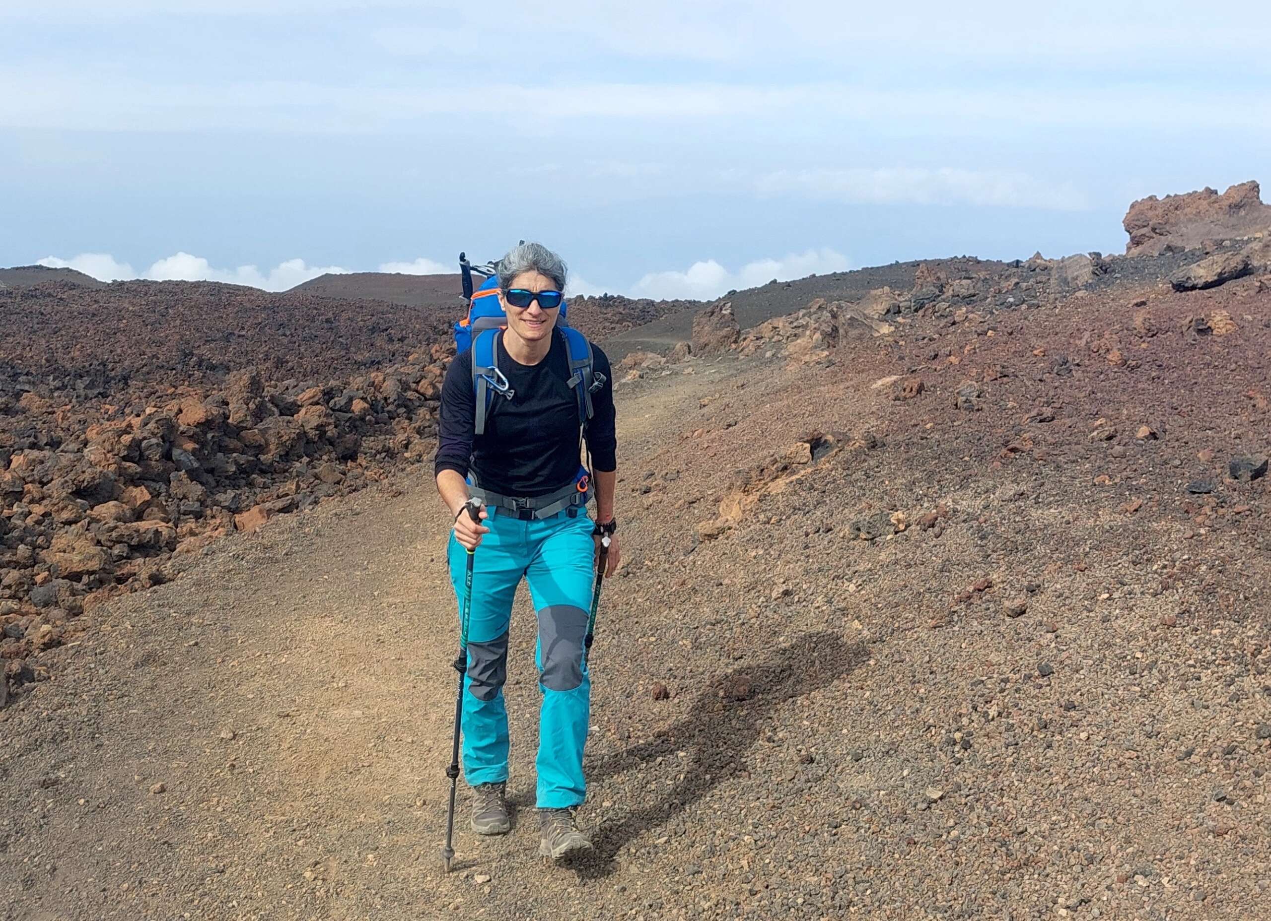 Cristina aparece en el centro de la foto de cuerpo entero practicando marcha nórdica. Está en un paraje desértico, en las cañadas del Teide. Viste ropa de montaña y porta una mochila.