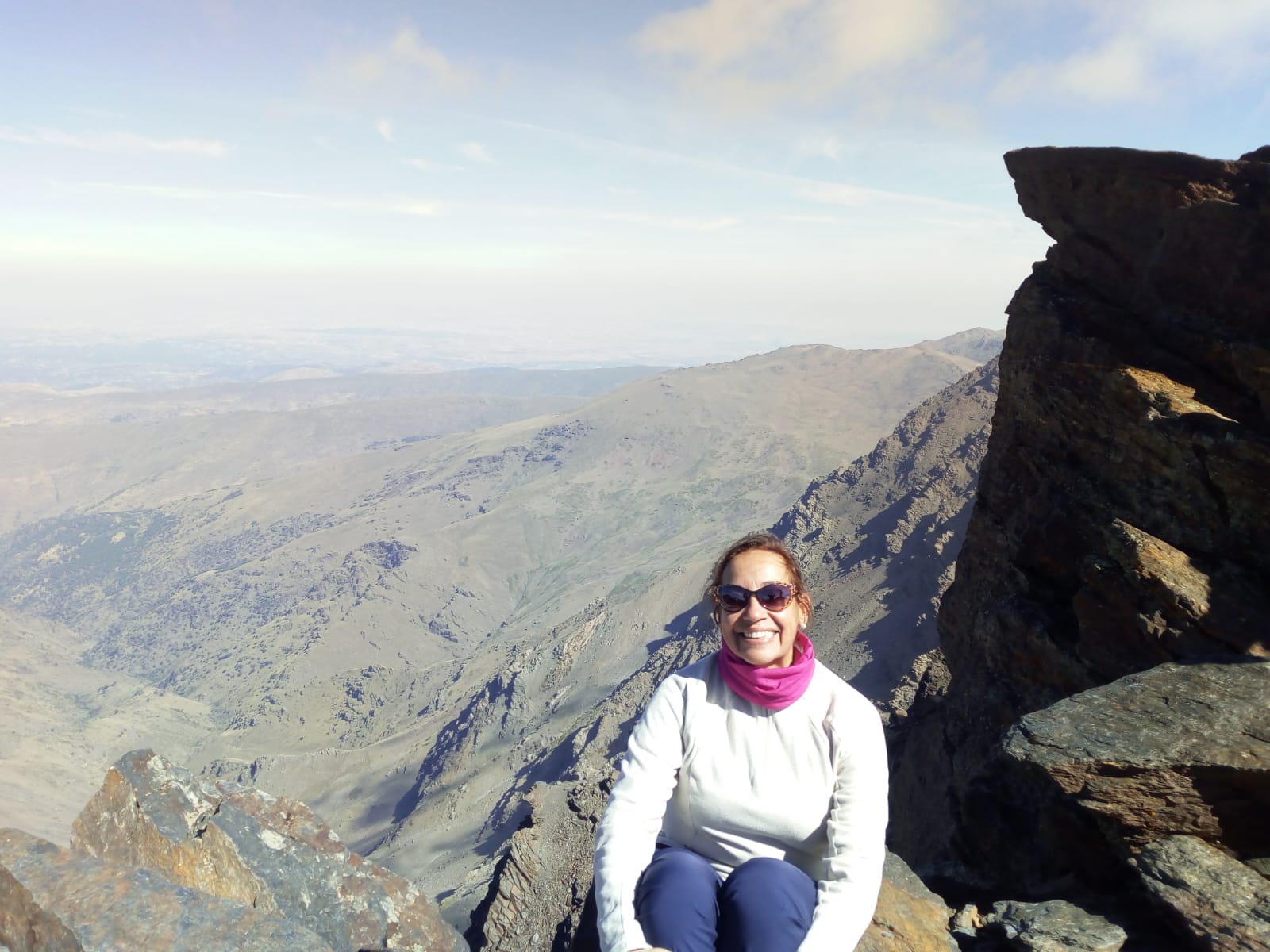 Chusa aparece sentada en una roca, en su primera subida al Mulhacén, que con sus 3718 metros es pico más alto de la península ibérica, al fondo un mar de nubes sobre Sierra Nevada. Lleva gafas de sol y un buff rosa al cuello. Tiene una gran sonrisa, ya que está feliz porque para ella está ascensión supuso algo muy importante.