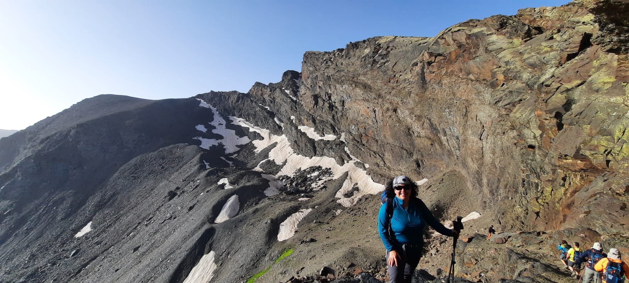 Magdalena aparece en primer plano, de pie, bajando desde Posiciones por el Veredón Superior a los Corrales del Veleta. Lleva una camiseta azul, gafas de sol, un pañuelo en su pelo rizado y unos bastones en su mano. Sonríe a la cámara, tranquila, feliz. De fondo la cara norte del Veleta, Sierra Nevada, junio del 2022