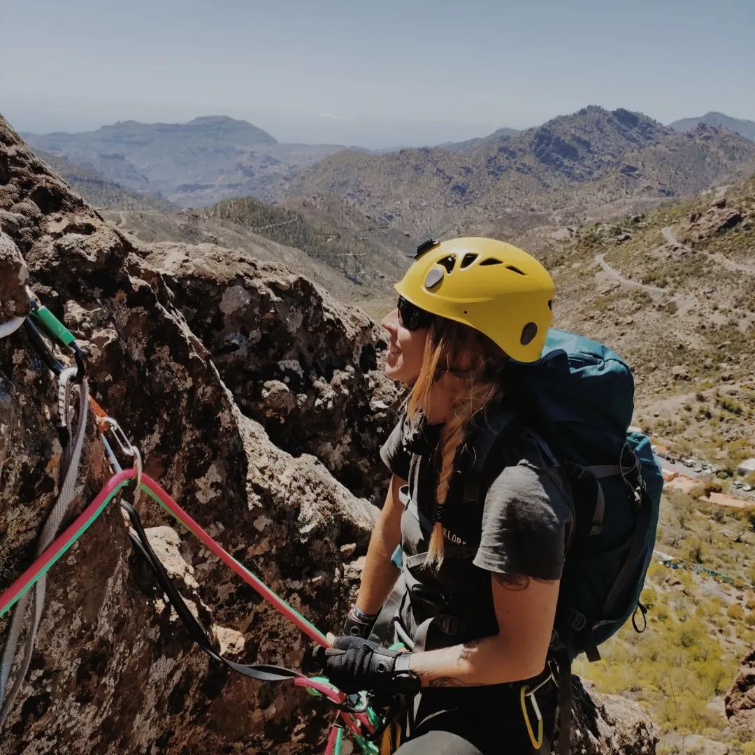 Descripción foto: Tania aparece en primer plano escalando, en el 2022, “Un paseo para disfrutarlo”, que es una vía de la cara Oeste de Roque Espinillo, Ayacata, Gran Canaria, abierta el 09 de diciembre de 1967. Está de perfil, sonriéndole a la roca, con gafas de sol, casco amarillo, trenza rubia y mochila azul a los hombros, en el horizonte siempre las montañas.