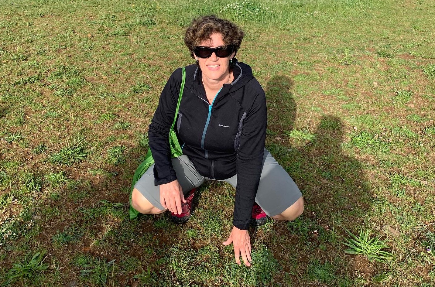 Begoña aparece en el centro de la foto en cuclillas y con una mano apoyada en la hierba. Se encuentra vestida con ropa de montaña, bermudas y forro polar.