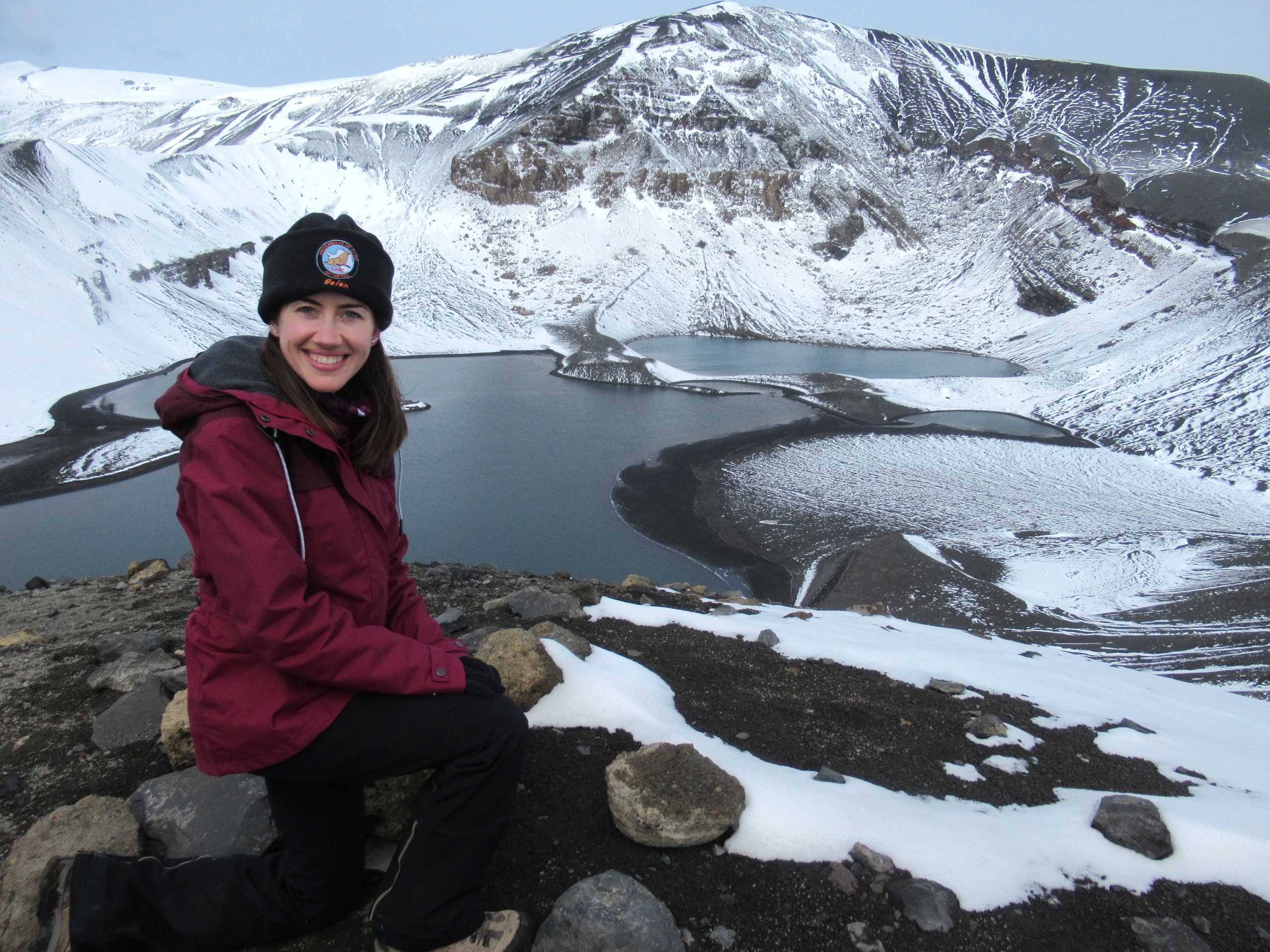 Belén aparece sonriente sentada en primer plano, sobre una piedra con ropa de abrigo y un gorro con el logo de su proyecto de la Universidad de Cádiz. Detrás un lago y una montaña nevada, llamados lago Irizar y monte Irizar. Esta fotografía está tomada en la isla Decepción, Antártida, desde Cerro Caliente.