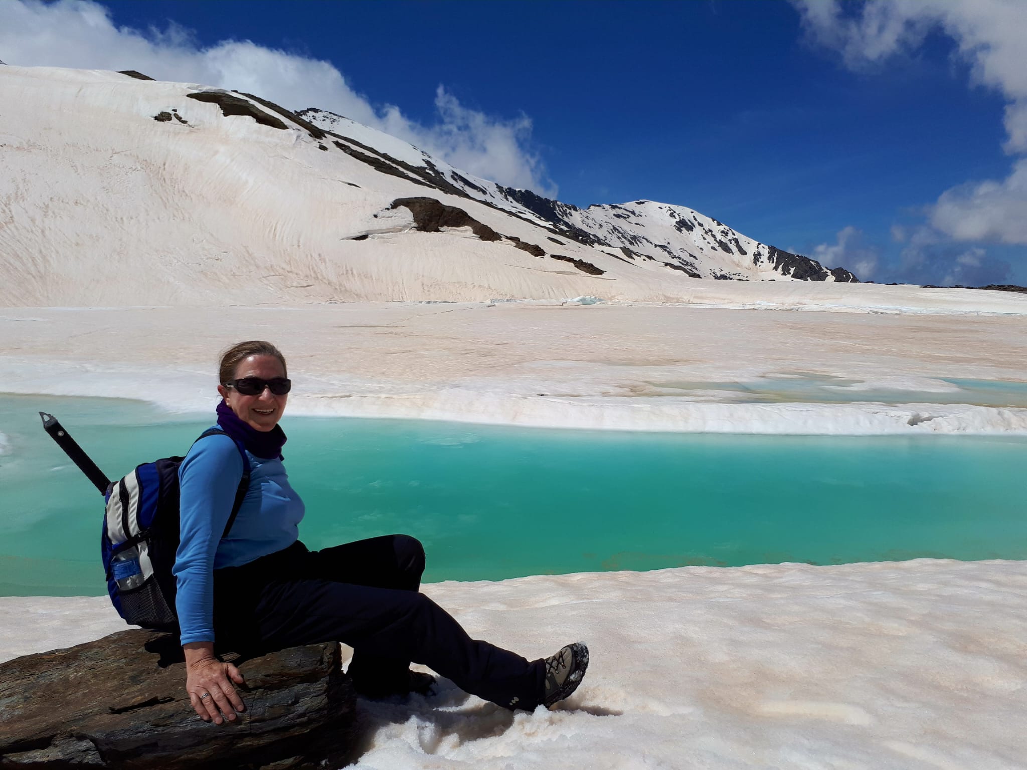 Ana aparece muy sonriente en la parte inferior izquierda de la foto sentada sobre una piedra en la orilla nevada de la Laguna de las Yeguas en Sierra Nevada. El paisaje está todo nevado, la misma laguna tiene una capa de nieve encima. Ana viste pantalón largo negro y camiseta de manga larga azul, así como botas de montaña, gafas de sol y una mochila en la que destaca su piolet.