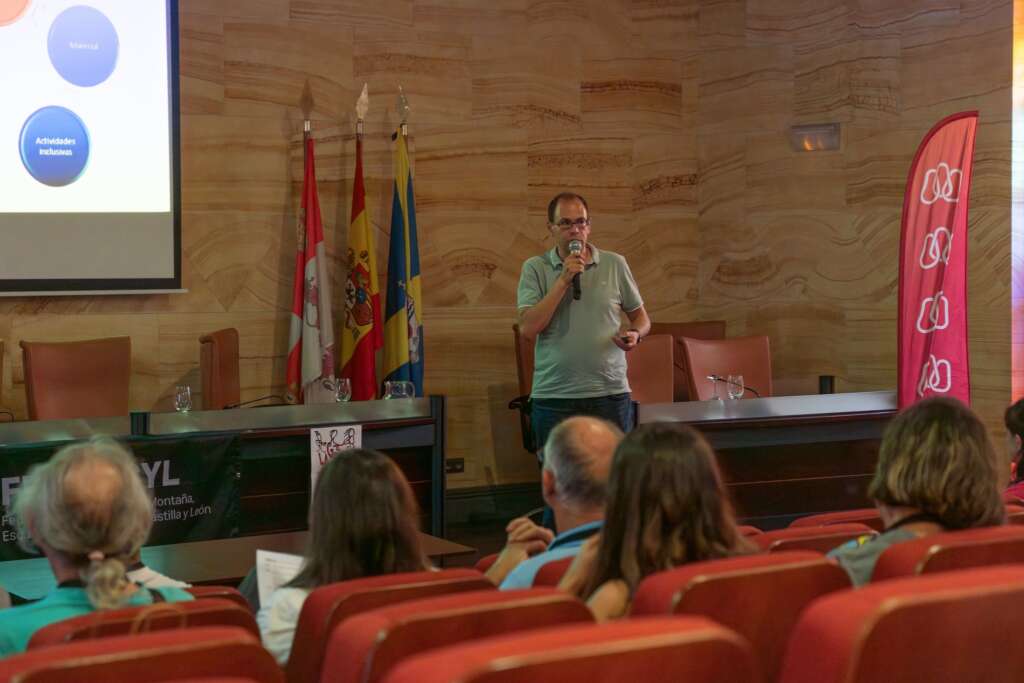 4. Aparece Javier González, Presidente de la Federación de Montaña de Castilla y León, de pie, con micrófono en la mano e impartiendo su ponencia: Pasado, presente y futuro de la accesibilidad de los itinerarios en la naturaleza