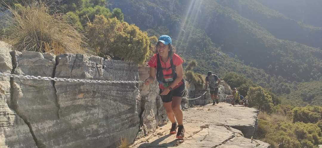 Charo está corriendo en una carrera por montaña, está agarrada a una cadena de seguridad, sonríe feliz, viste camiseta roja, mochila ligera y una gorra azul.