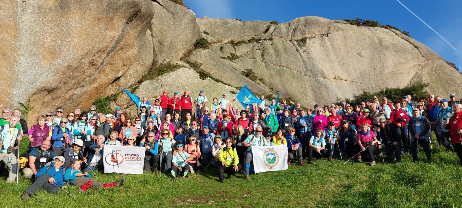 Un nutrido y vivaz grupo de Deportistas con Veteranía con las banderas de sus clubes, en la pradera sur de la base del Pico da Frouxeira, detrás el canchal granítico del Pico con un cielo azul intenso, en la 48 Marcha Anual de Deportistas con Veteranía del Centenario de la FEDME 2022.