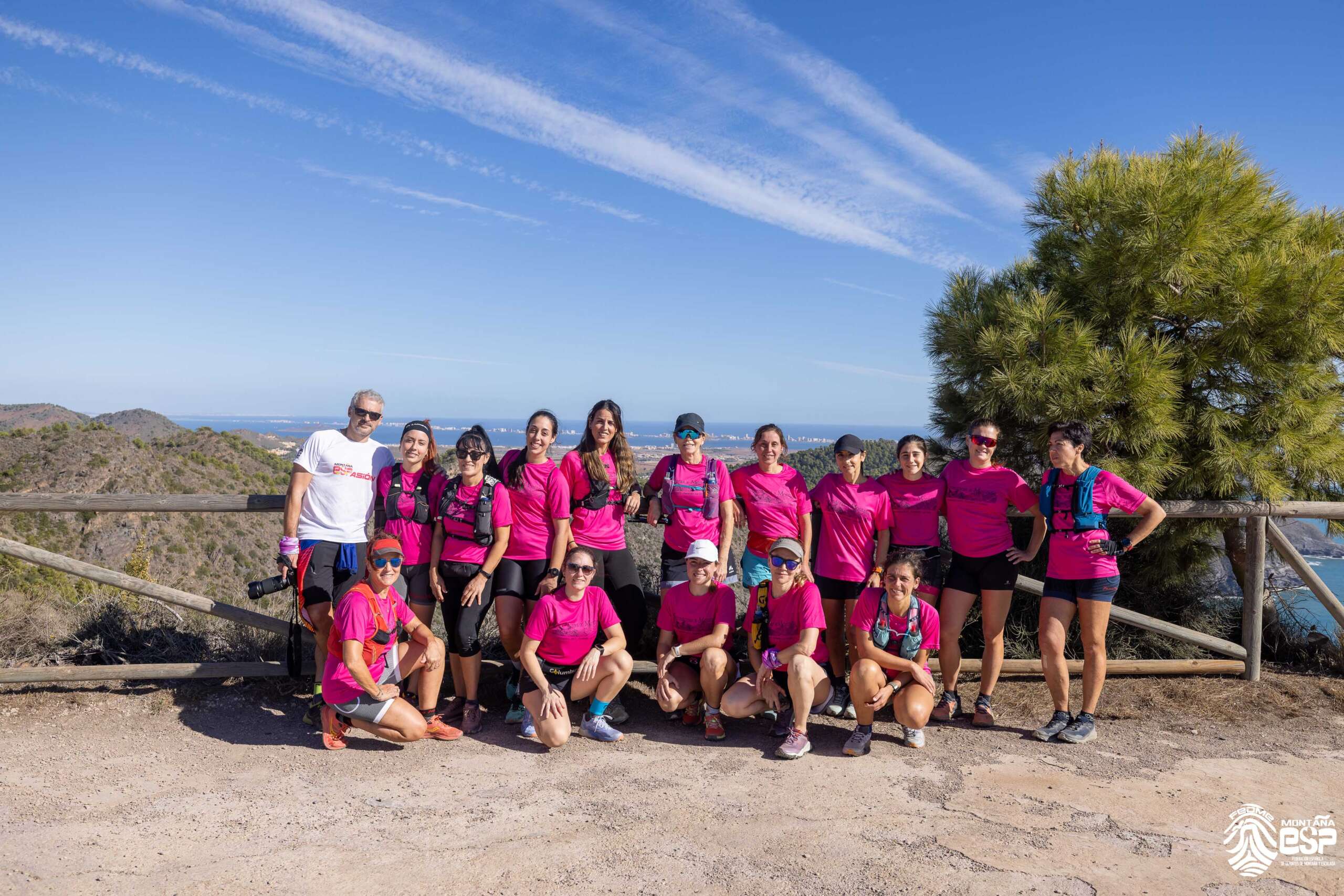 En primer plano aparecen todas las participantes al Encuentro equipadas con camiseta rosa del evento y pantalón corto. Unas están de pie y otras en cuclillas delante. Tras ellas un paisaje de montaña y mar con cielo azul intenso