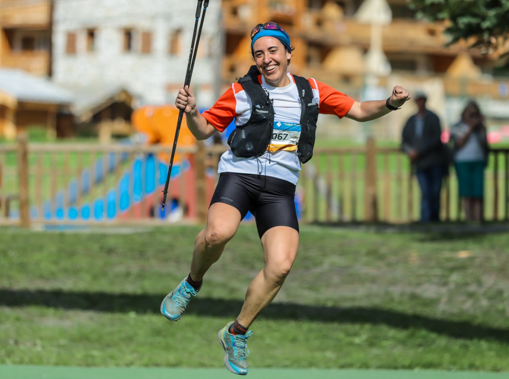 Elvira está llegando a meta en una carrera por montaña, está saltando en el aire, con los bastones en su mano derecha. Luce una preciosa sonrisa de satisfacción. Va vestida con unas mallas negras, una camiseta blanca y azul y una mochila de correr sobre los hombros.