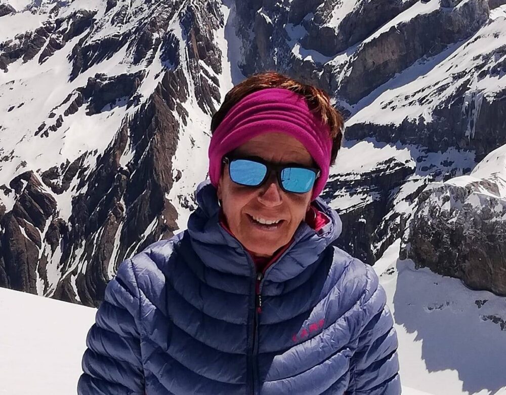 Mónica Vergé aparece en primer plano, con chaqueta de pluma azul, gafas de sol y cinta rosa en el pelo. Tras ella una pared de roca y hielo muy abrupta