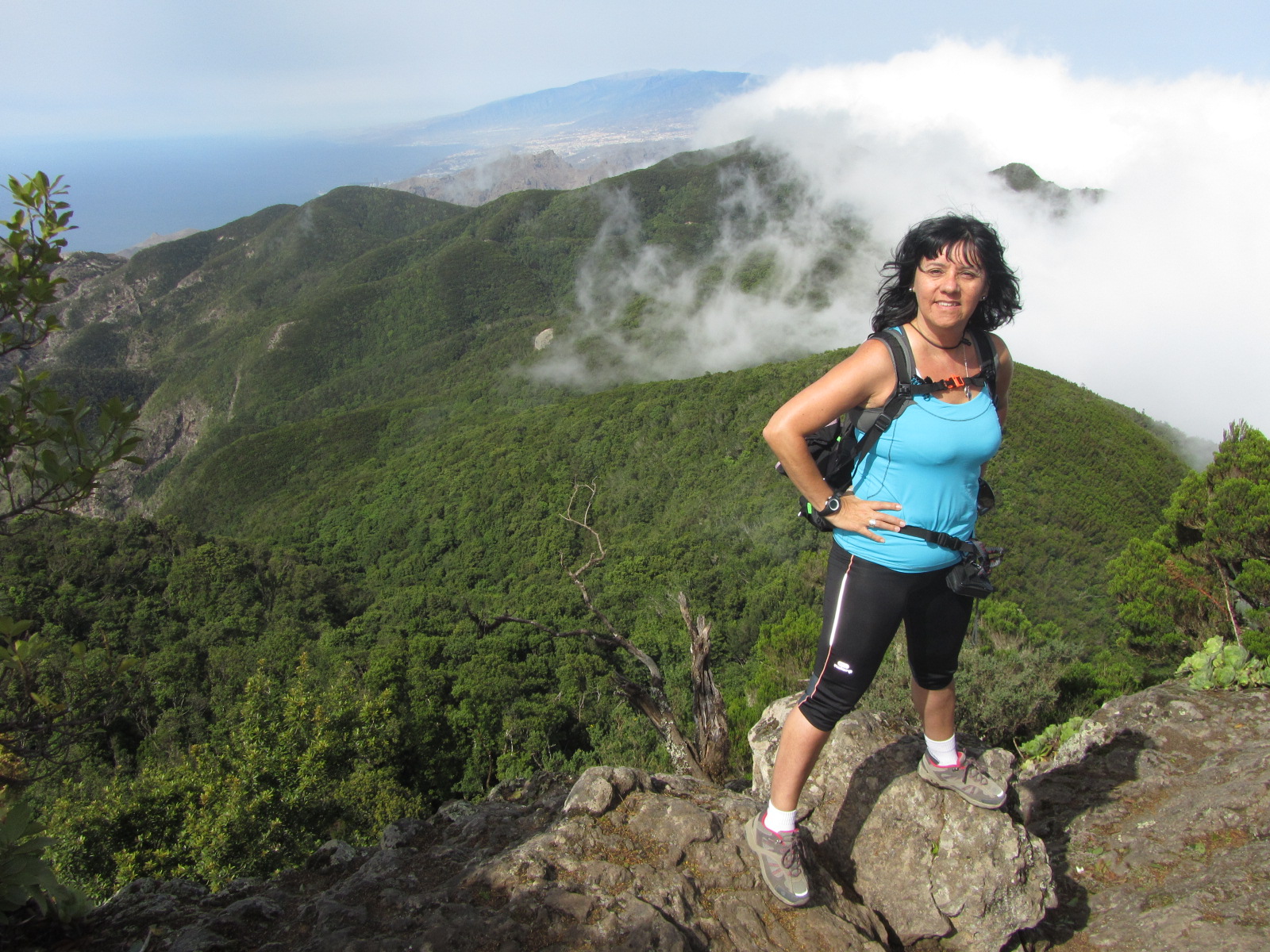 Cande Rodríguez Izquierdo aparece en primer plano de pie sobre una piedra, con las manos a la cintura, con malla negra y camiseta de tirantes azul y sonriente. Tras ella un espléndido paisaje verde de El Pijaral perteneciente al Parque Rural de Anaga en Tenerife y al fondo el mar