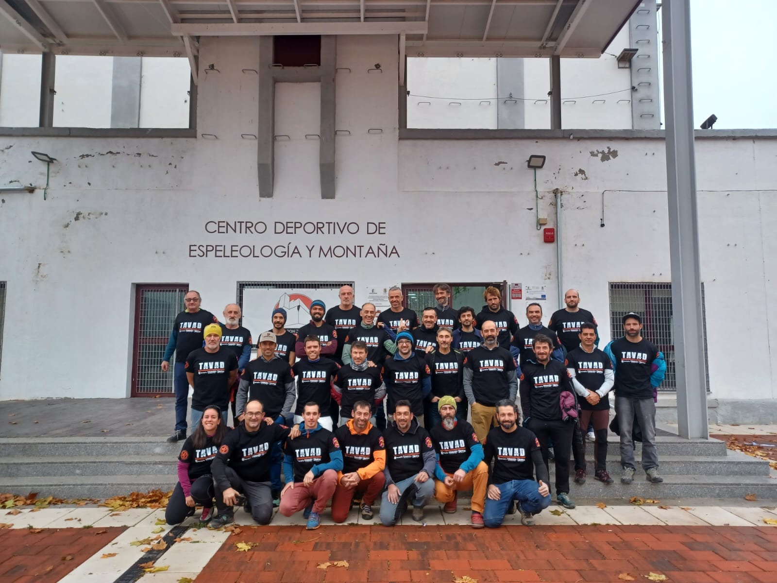 : en la foto aparece un grupo de 30 personas, todas ellas vestidas con equipación deportiva y con una camiseta negra en la que está escrito TAVAD con letras mayúsculas blancas. El grupo está colocado en cuatro filas y subidos en los escalones externos de entrada al Silo de Chillarón: Centro Deportivo de Espeleología y Montaña.
