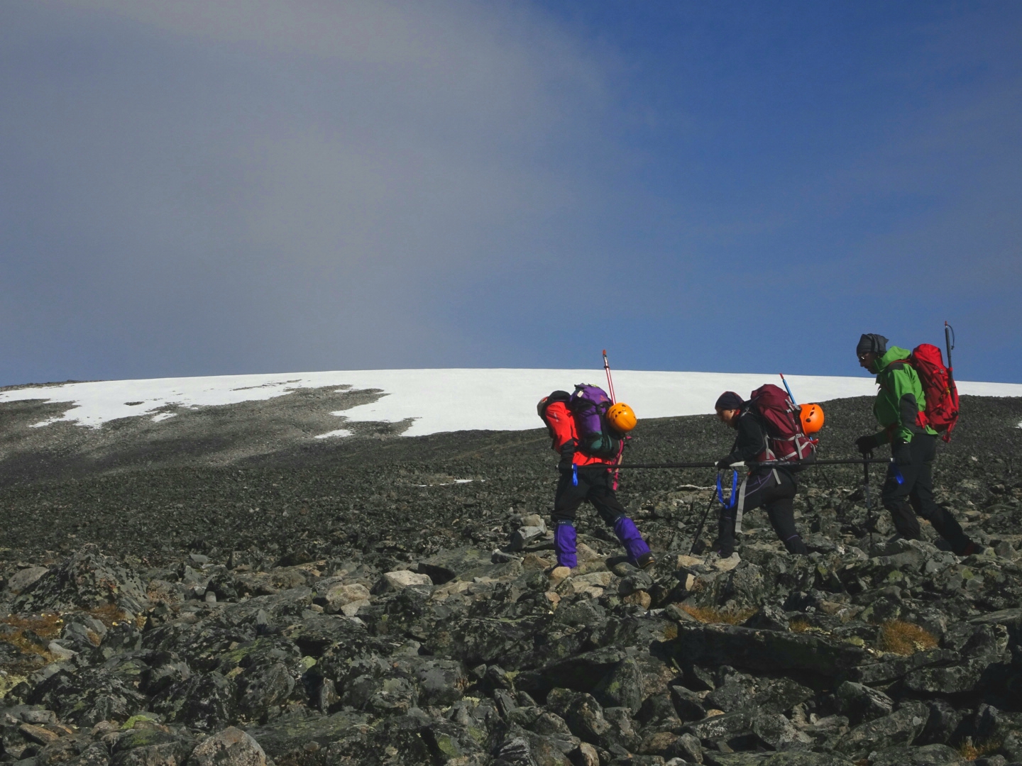 En primer plano se ven tres deportistas caminando por una zona empedrada de montaña. La primera y última persona portan una barra direccional como guías y en medio la tercera persona montaña con discapacidad visual. Van equipados de alta montaña con mochilas y en ellas casco y piolet. Se encaminan hacia zona de nieve o hielo que se ve al fondo