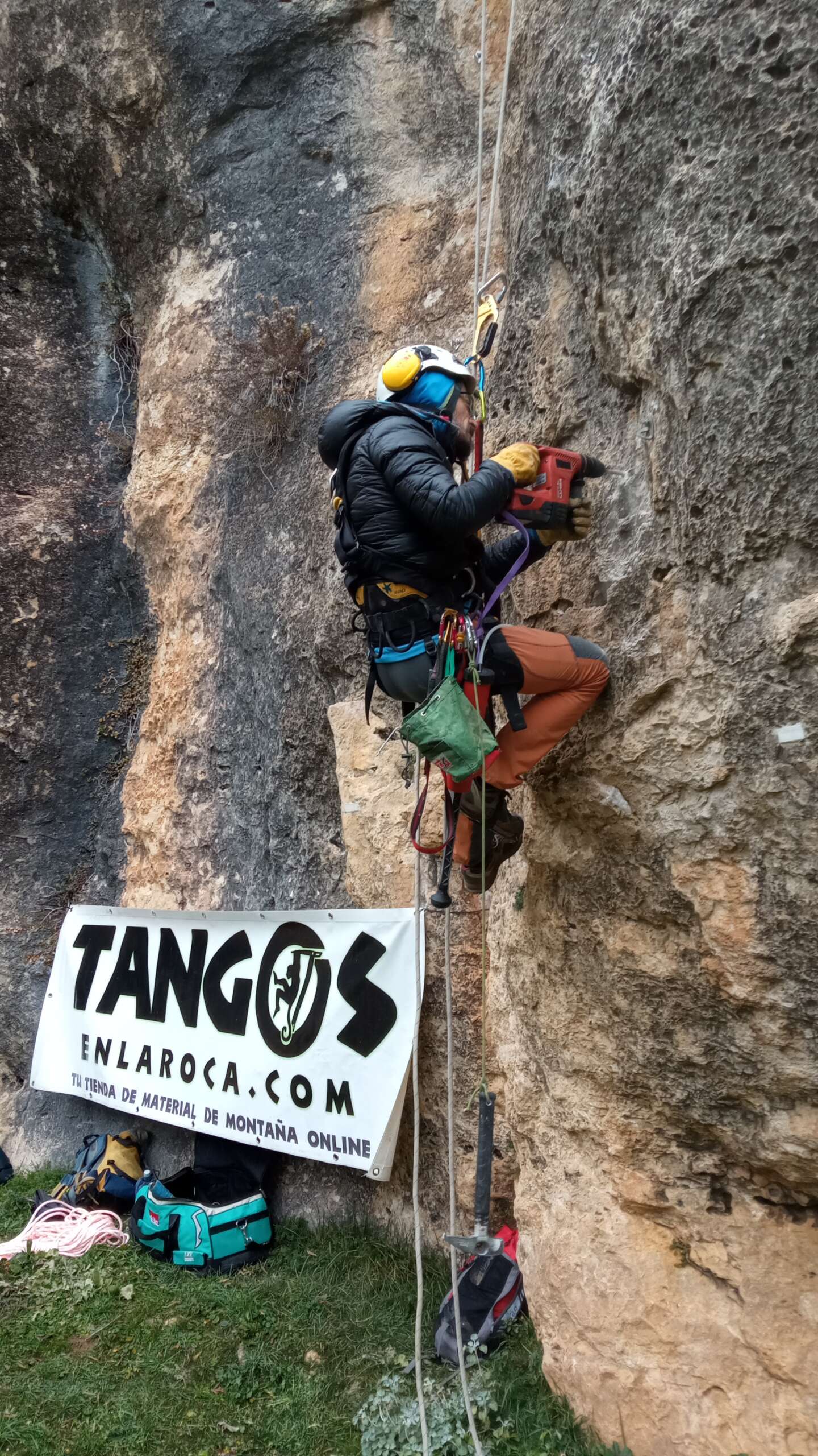 Un escalador en roca equipando una via. Con la imagen de una pancarta publicitaria de Tanogs en la Roca