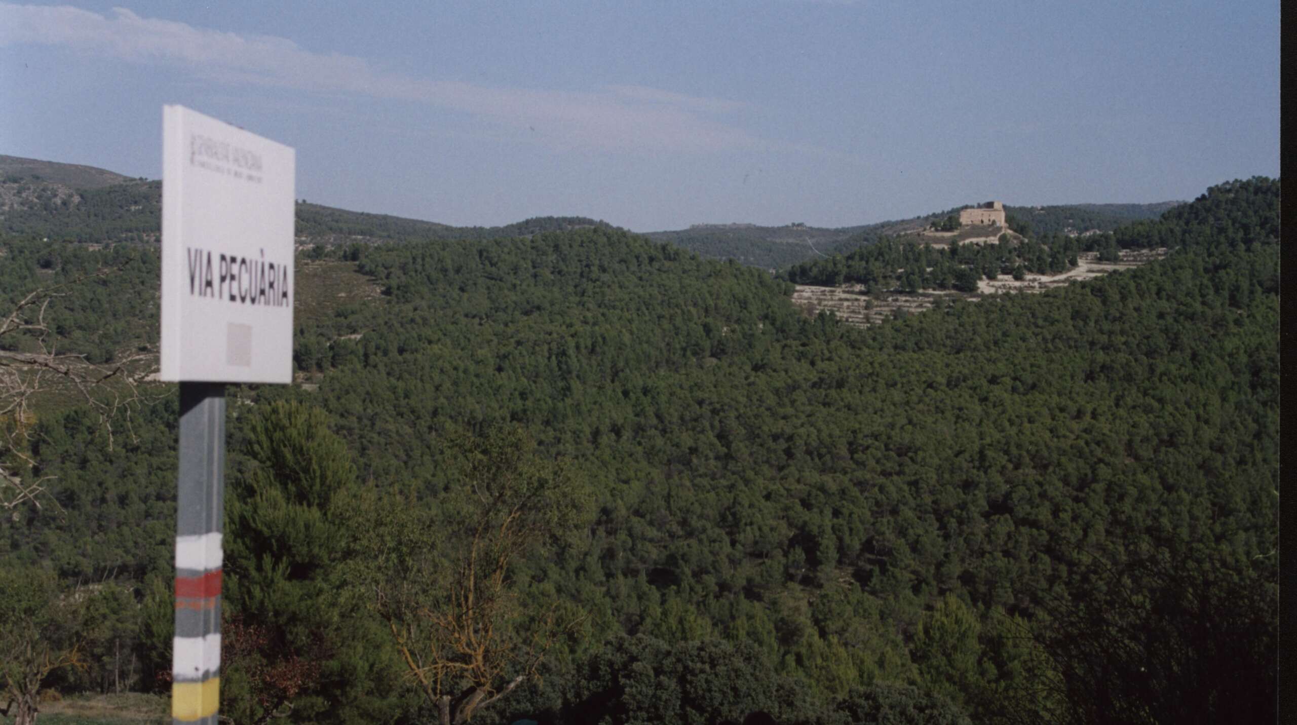 La imagen es una fotografía de un paisaje de baja montaña con cielo de color azul y algunas nubes. Al fondo se pueden observar una construcción en lo alto de una colina y, en primer plano a la izquierda, hay una seña metálica de “VIA PECUARIA”. En el poste de la señal están pintadas las marcas de Sendero GR (línea blanca horizontal y debajo otra roja) y de Sendero PR (línea blanca horizontal y debajo otra amarilla).