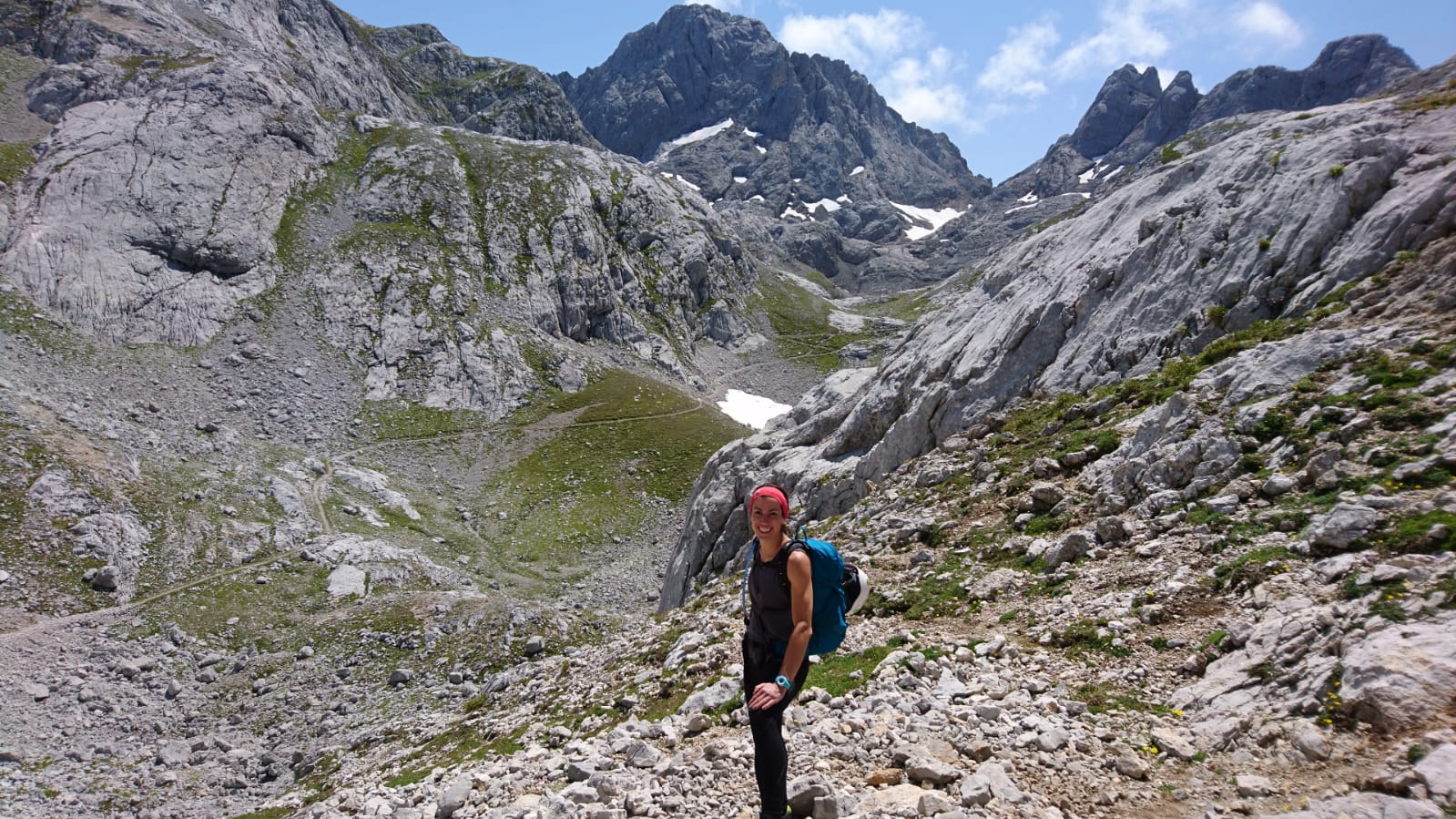 Noelia aparece en primer plano durante una jornada de senderismo en el Parque Nacional de los Picos de Europa. Va vestida con pantalón y camiseta de color negro, una cinta roja en su pelo y mochila en la espalda. Tras ella un impresionante paisaje de Picos, con roca, nieve y vegetación verde