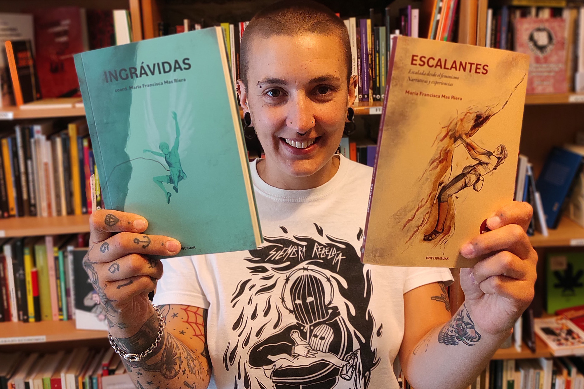 MF aparece en el centro de la foto de cintura para arriba mostrando sus libros en cada mano. Al fondo una estantería repleta de libros en el local de la DDT en Bilbao.