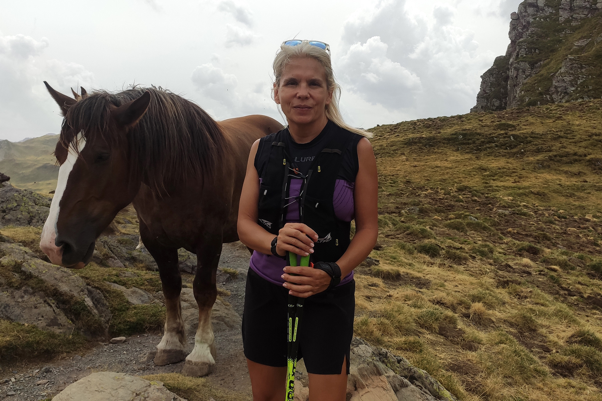 Peli aparece en medio de la foto en un paisaje montañoso con ropa de montaña y bastones de marcha nórdica. A su izquierda y justo detrás tiene un caballo que parece estar posando junto a ella.