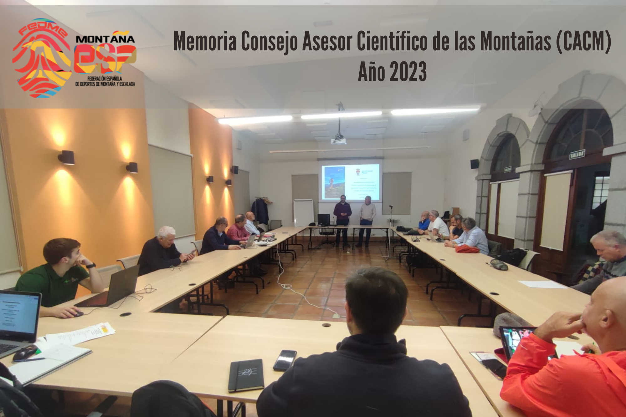 fotografía de un grupo de personas sentadas reunidas en una sala cuyas mesas están dispuestas en forma de U. Atienden a dos personas situadas en delante de una pantalla en el extremo abierto de las mesas. Sobre la imagen, en el borde superior hay un rectángulo de color gris transparente donde están incluidos el logo de FEDME en la esquina izquierda (una huella dactilar de colores rojo, amarillo y azul cuyas líneas forman el Torreón de los Galayos con un pájaro sobrevolándolo. A la derecha de la huella aparecen las letras Montaña en negro, debajo ESP en colores rojo y amarillo y debajo las letras Federación Española de Deportes de Montaña y Escalada en negro) y en el centro-derecha está escrito: “Memoria Consejo Asesor Científico de las Montañas (CACM) Año 2023”.