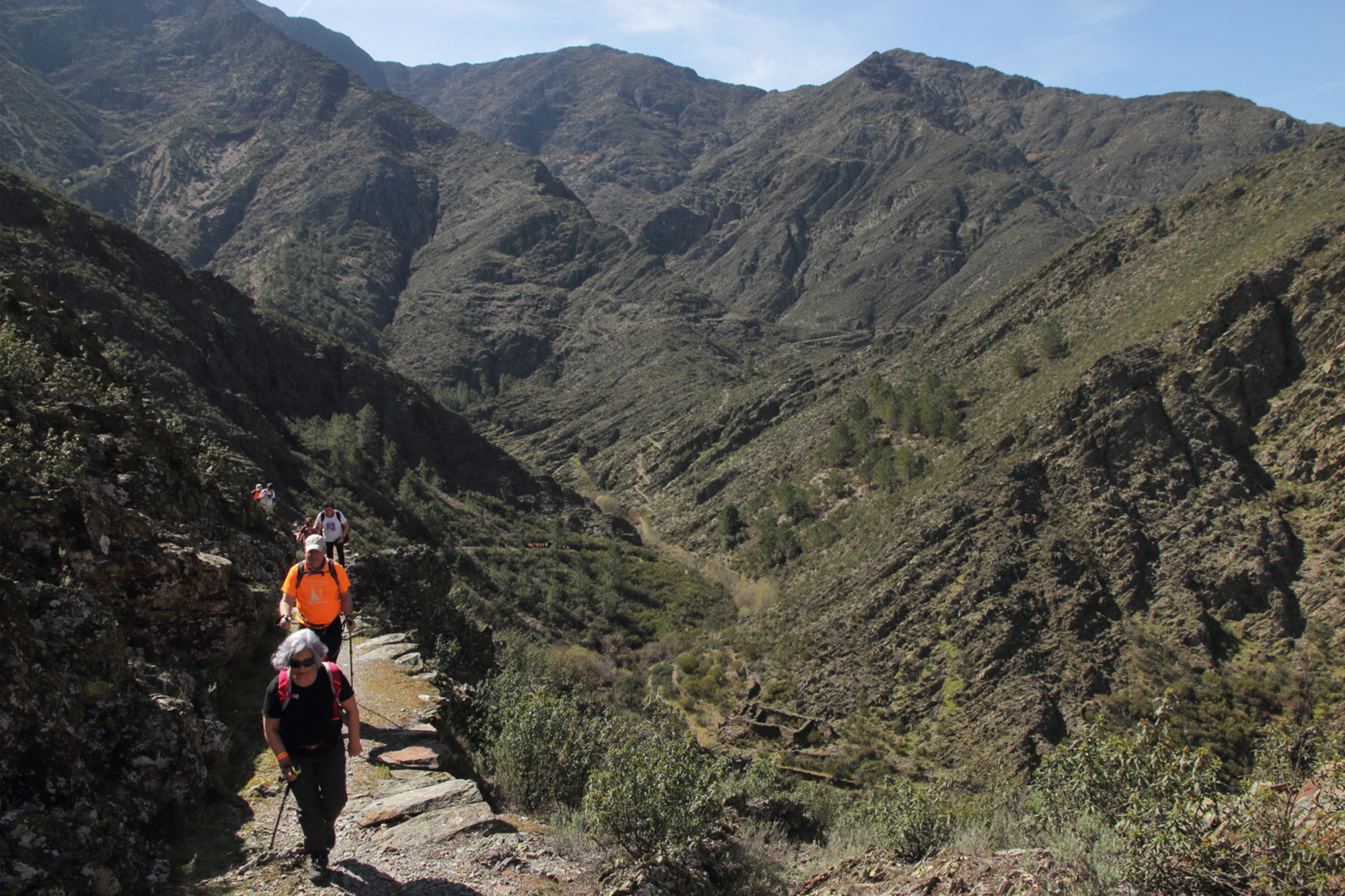 Foto en color que presenta a un grupo de senderistas ascendiendo por un camino antiguo, abierto en una ladera de un valle montañoso, en el norte de Extremadura