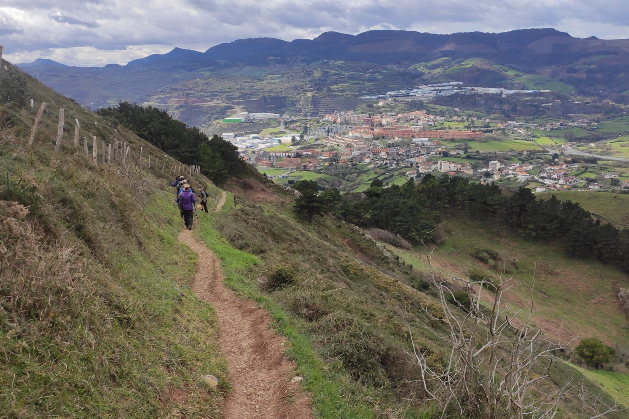 Foto en color que presenta a un grupo de senderistas caminando sobre una estrecha senda, trazada en una ladera herbácea. En el fondo del valle aparece una población.