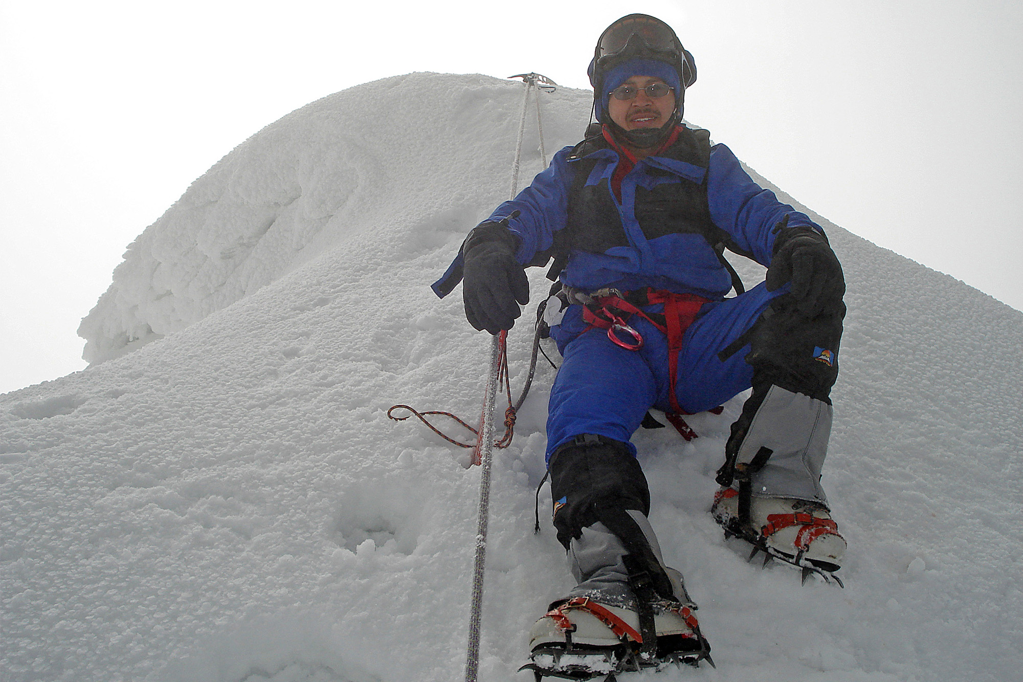 Christian Rodríguez en primer plano, sonriendo a la cámara, sentado en un montículo de hielo, mientras se asegura con una cuerda y varios piolets. Volcán Chimborazo de 6300 mts. Ecuador.