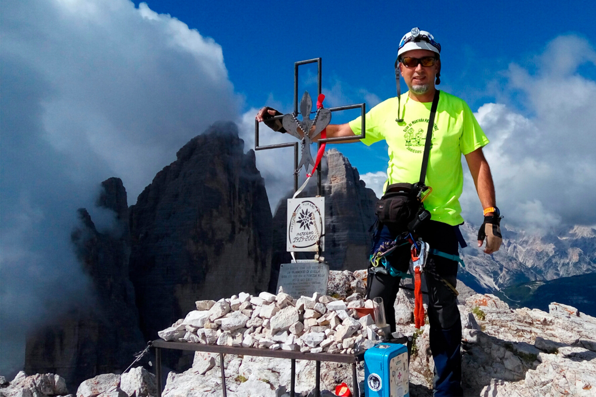En la foto estoy tocando una cruz de forja con el símbolo de la flor de lis en el centro, en la cima de una montaña de los Dolomitas. Al fondo se ven las famosas Tres Cimas de Lavaredo