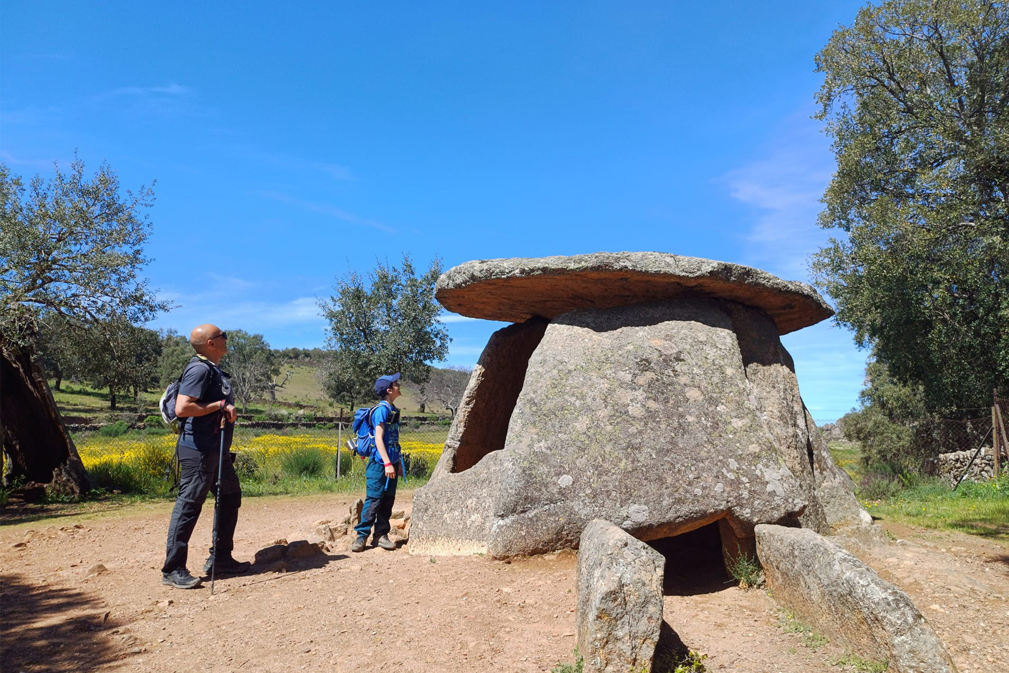 Ilustración a color que presenta a dos senderistas ante el dolmen de los Mellizos (Valencia de Alcántara-Cáceres), ubicado en una ruta de senderismo del Circuito Camina Extremadura, Liga Ibérica de Senderismo.