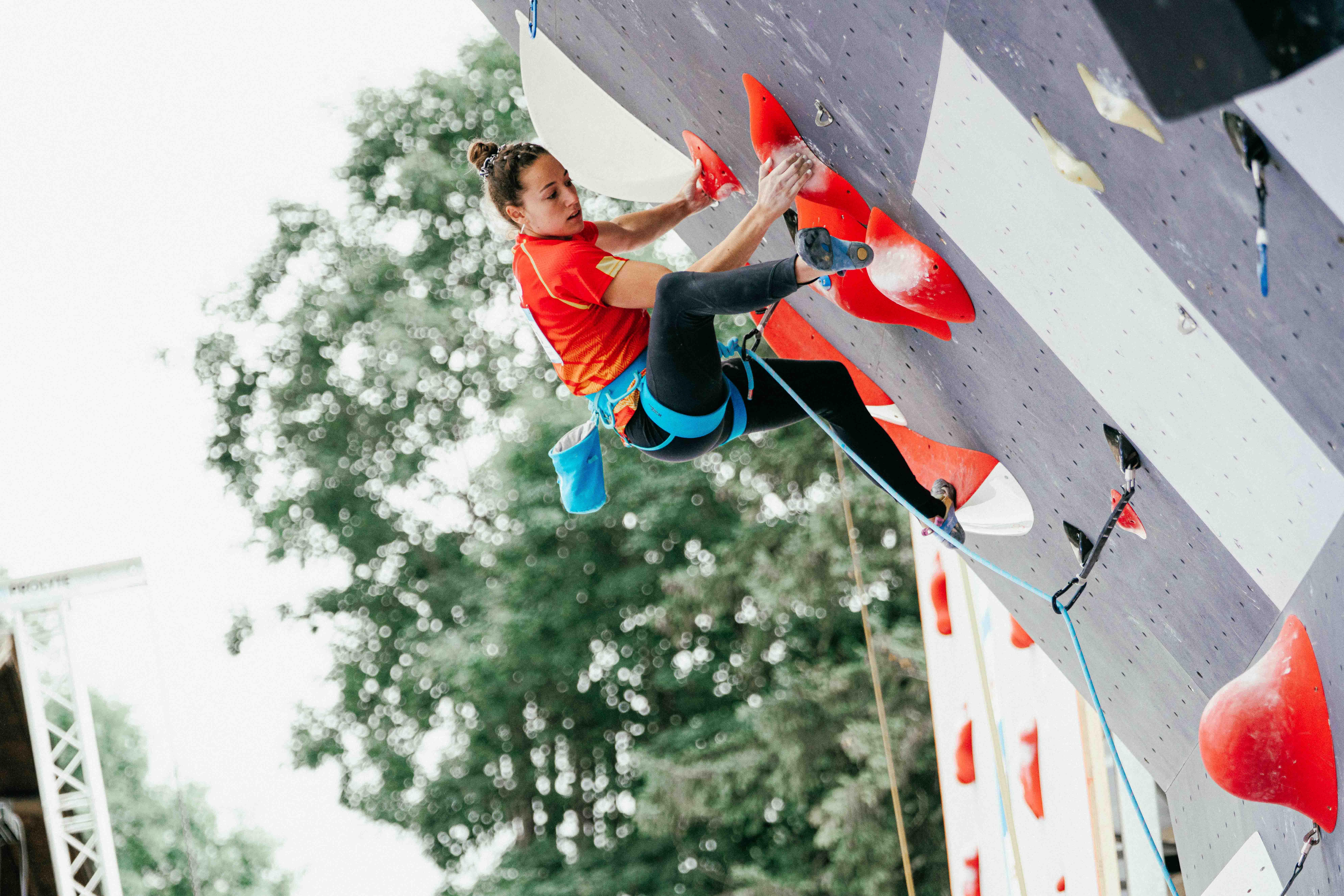 Aida está escalando en rocódromo, en prueba competitiva de escalada. Lleva camiseta roja de la selección española y malla negra. Se encuentra realizando un movimiento complicado de taloneo en una de las presas rojas
