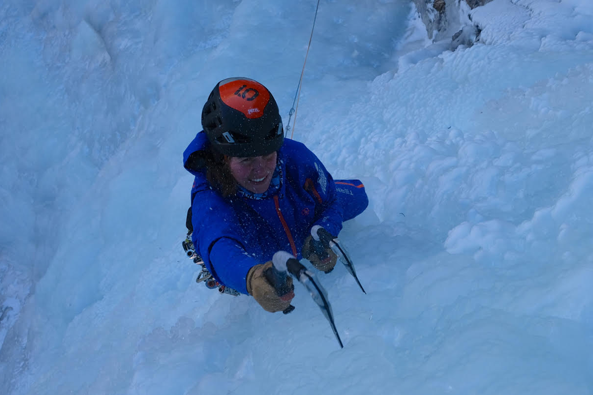 Marina escalando en hielo en el macizo de Ecrins, Francia, equipada con ropa azul y casco rojo, con los dos pioletes agarrados y clavados en el hielo en primer plano, ante una impresionante pared de hielo.