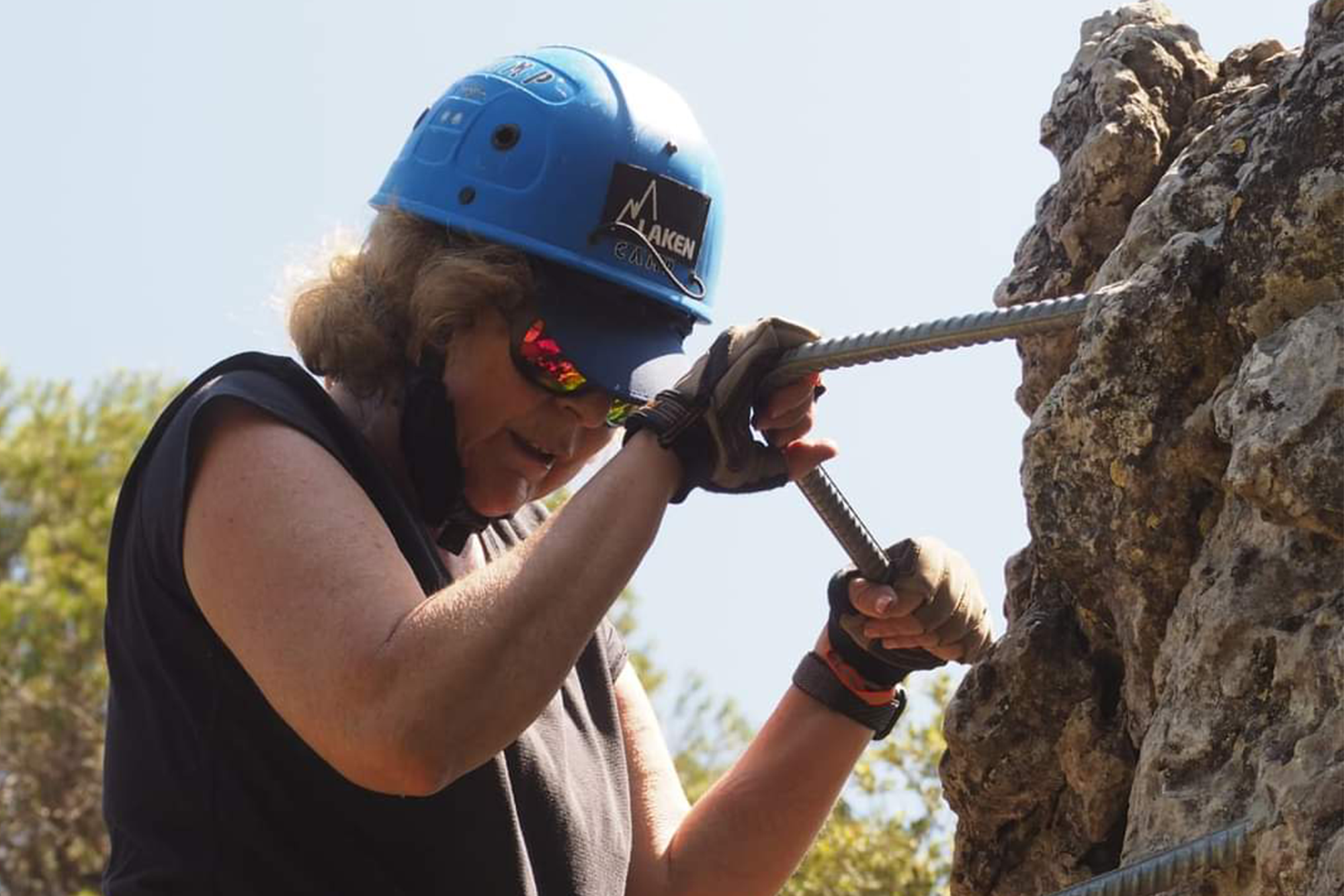 Foto tomada en la vía Ferrata de Priego en verano del 2023 a la que volví 6 años después de mi Parkinson. Una mujer con casco azul está escalando una roca. Lleva una camiseta sin mangas negra y pantalones rojos. Tiene un arnés de seguridad y está sujetando una cuerda de escalada. Hay un mosquetón y una botella de agua verde colgando de su arnés. Un día soleado y despejado.