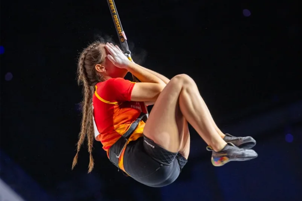 María aparece colgada de cuerda de escalada en competición de velocidad, con rodillas encogidas y las manos tapándose la cara