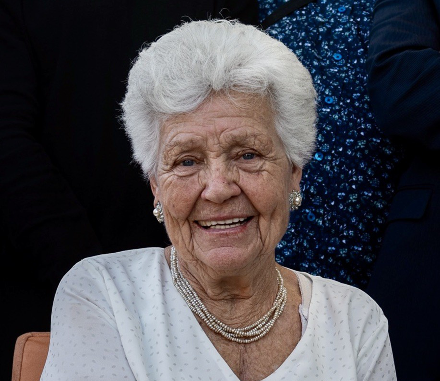 Palmira aparece en primer plano muy sonriente, con camisa blanca a juego con su pelo corto y cano y con bonitos pendientes plateados así como el collar de su cuello
